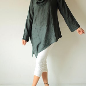May include: A dark green, long-sleeved, asymmetrical tunic top with a cowl neck. The top is made of a soft, drapey fabric and has a relaxed fit. The model is wearing white pants and black sandals.