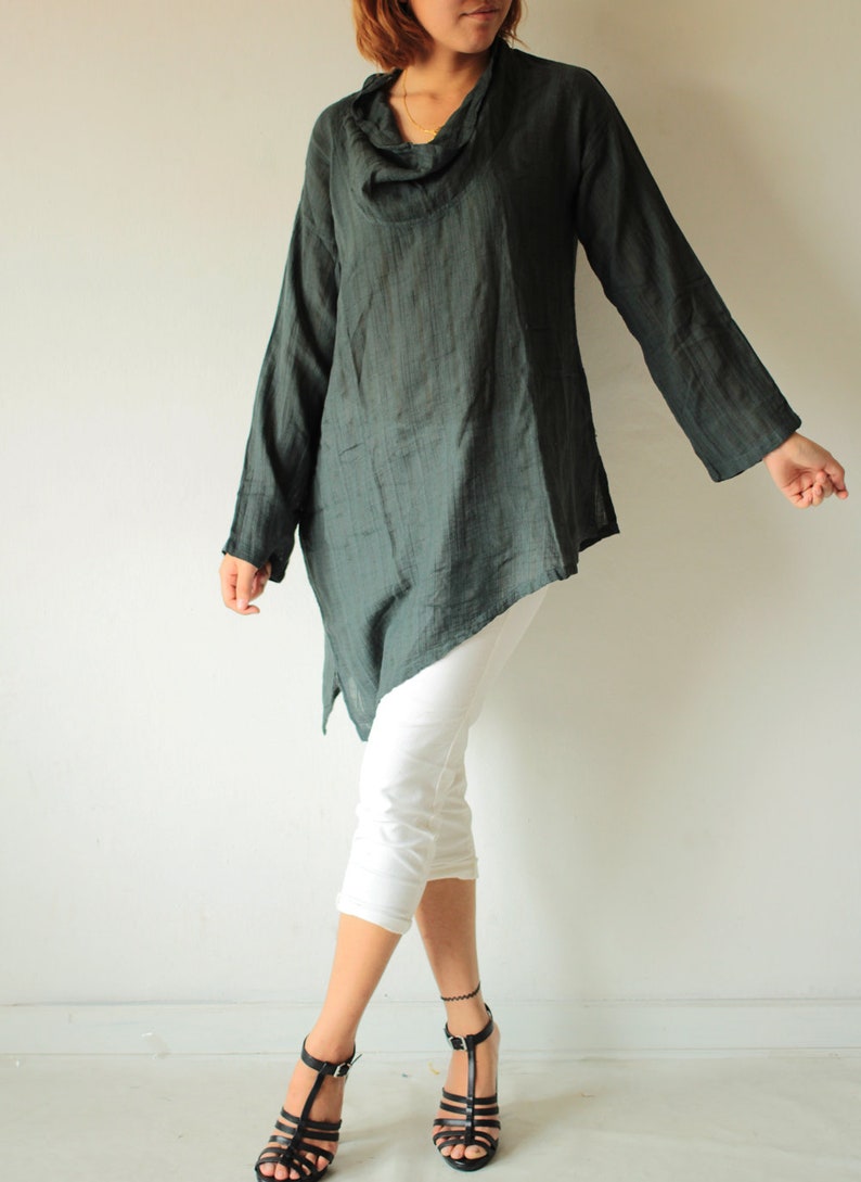May include: A dark green, long-sleeved, asymmetrical tunic top with a cowl neck. The top is made of a soft, drapey fabric and has a relaxed fit. The model is wearing white pants and black sandals.