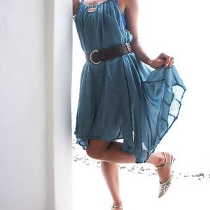 May include: A woman wearing a light blue sleeveless dress with a brown woven belt and silver sandals. The dress has a flowing, loose fit and is cinched at the waist with the belt.