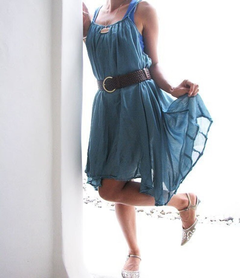 May include: A woman wearing a light blue sleeveless dress with a brown woven belt and silver sandals. The dress has a flowing, loose fit and is cinched at the waist with the belt.