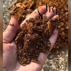May include: A close-up of a hand holding a quantity of brown reptile substrate, possibly wood chips. The material is coarse and textured. The image includes a logo that says "THE REPTILE JUNGLE" in a circle.