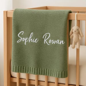 May include: A sage green knitted baby blanket with the name "Sophie Rowan" embroidered in white script. A small beige bunny toy hangs from the wooden crib. The blanket is folded over the crib rail.