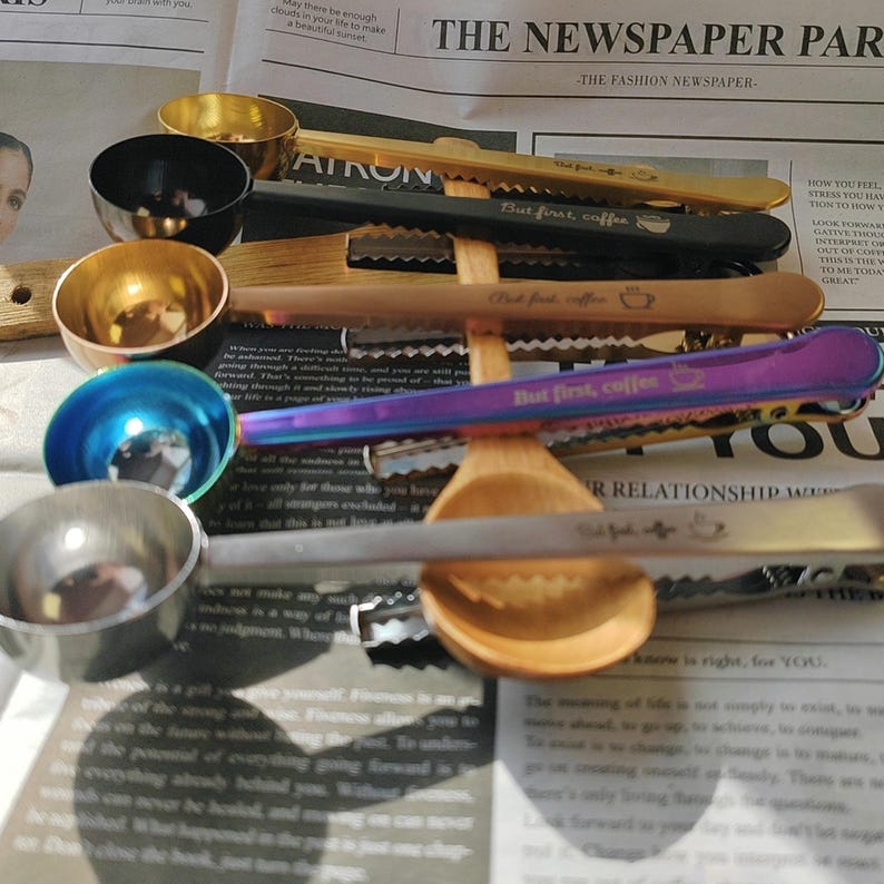 May include: A set of six colorful metal measuring spoons with a wooden spoon and a set of four metal hair clips. The hair clips are engraved with the text "But first, coffee".