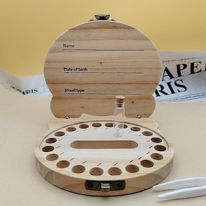 Op de afbeelding: Houten bewaardoos voor babytanden met een scharnierend deksel. Het deksel heeft lijnen voor naam, geboortedatum en bloedgroep. De doos heeft een cirkelvormige basis met gaten voor tanden en een klein glazen flesje.