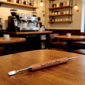 May include: A coffee scoop and tamper tool with a wooden handle and the phrase "but first coffee" engraved on it. The tool has a scoop on one end and a pointed tamper on the other. The tool is lying on a wooden table.