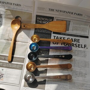 May include: A set of six coffee measuring spoons with wooden handles. The spoons are silver with gold accents and have inspirational quotes engraved on them. The spoons are arranged on a newspaper with the words "Take Care of Yourself" printed on it.