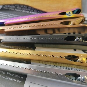 May include: Five hair clips in different colors, including black, gold, silver, and pink. The clips have a zig-zag pattern on the top and a spring mechanism on the bottom.
