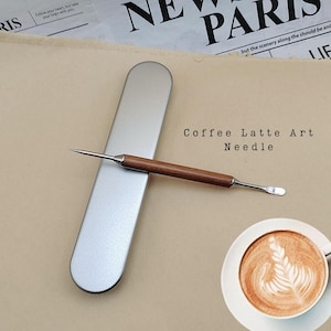 May include: A coffee latte art needle with a wooden handle and a stainless steel tip and spoon end. The needle is next to a silver metal case and a cup of coffee with latte art. The text "Coffee Latte Art Needle" is visible.