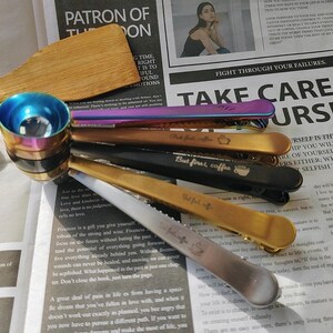 May include: A set of six gold, silver, and black metal hair clips with engraved text, including "But first, coffee" and "The best is yet to come". The clips are arranged on a newspaper with a wooden spoon and a rainbow-colored measuring spoon.