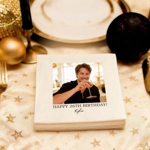 Op de afbeelding: Een stapel witte papieren servetten met een afgedrukte foto van een lachende persoon die een wijnglas vasthoudt. De tekst "HAPPY 26TH BIRTHDAY! Kyle" staat eronder. De servetten liggen op een gouden tafelkleed met sterrenpatroon.