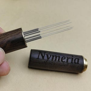 Op de afbeelding: Een donkerhouten container met het woord "Nymeria" gegraveerd op de zijkant, met daarin verschillende acupunctuurnaalden. De naalden zijn zilverkleurig en gebundeld. De container is open en onthult de naalden.