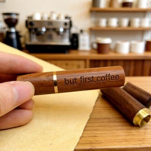 May include: A wooden coffee accessory with the engraved text "but first coffee". The accessory is cylindrical with a gold band and a gold-colored end. The background shows a coffee shop setting with coffee machines and cups.