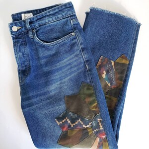 May include: Blue denim jeans with a decorative floral design on the lower leg. The floral design incorporates various patterned fabrics in shades of brown, green, and blue. The jeans have a button and zip closure and a raw hem.