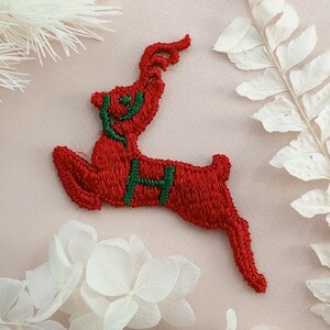 May include: Red felt reindeer applique with green embroidered details. The reindeer is in a leaping pose with its antlers raised.