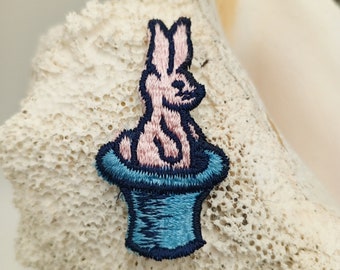 May include: A pink embroidered bunny rabbit sitting in a blue top hat. The hat has a black brim and a white lining.