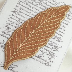 May include: A brown suede leaf-shaped patch with gold stitching. The patch is embroidered with a detailed leaf design.