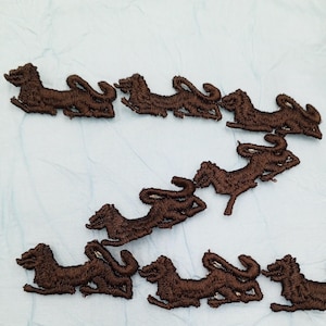 May include: A collection of brown embroidered patches in the shape of a running lion. The patches are arranged on a light blue fabric. The lion patches are detailed with embroidered features.