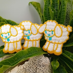 May include: Three white embroidered iron-on patches of cartoon lambs with yellow outlines and turquoise eyes. The patches are on a green leafy background.