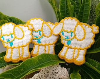 May include: Three white embroidered iron-on patches of cartoon lambs with yellow outlines and turquoise eyes. The patches are on a green leafy background.