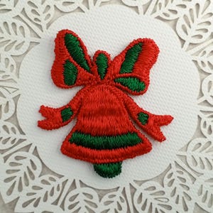 May include: Red and green embroidered Christmas bell with a bow. The bell is decorated with green stripes and the bow is green with red stripes.