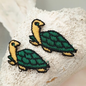 May include: Two green and yellow embroidered turtle patches. The turtles have a green shell with a grid pattern and a yellow head and legs.