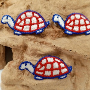 May include: Three embroidered turtle patches. Each turtle has a red and white shell with a blue outline and blue head and legs. The patches are on a textured, tan surface.