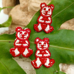 May include: Three embroidered patches in the shape of teddy bears. The bears are red with white accents, including the face, chest, and feet. The patches are arranged on a large green leaf.