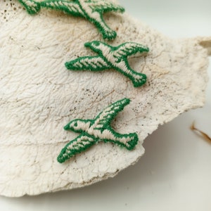 May include: Three green embroidered birds on a white textured surface. The birds are stitched with white thread and have a simple design.