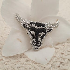May include: Black and silver embroidered bull head patch. The bull's head is facing forward with horns pointing up. The patch is on a white background.