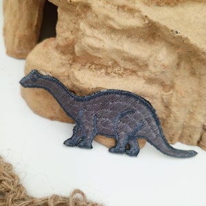 May include: A gray embroidered dinosaur patch with a long neck and tail. The patch is on a brown textured surface.