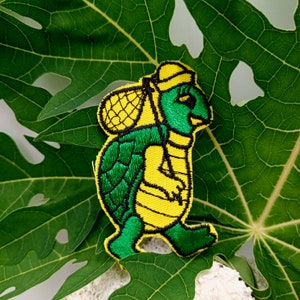 May include: A green and yellow embroidered patch of a turtle carrying a backpack. The turtle is wearing a hat and has a friendly expression.