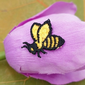 May include: A yellow and black embroidered bee applique on a purple fabric background.