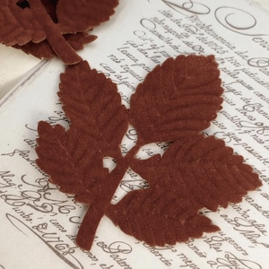 May include: A brown felt leaf sprig with five leaves and a stem. The leaves are cut out in a delicate pattern and have a velvety texture.