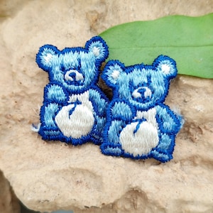 May include: Two blue embroidered teddy bear patches with white faces and white bellies. The bears are facing forward and have a simple design.