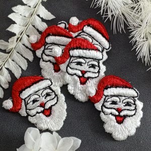 May include: Five white felt embroidered Santa Claus faces with red hats. The Santa faces have white beards and black eyes, noses, and mouths.