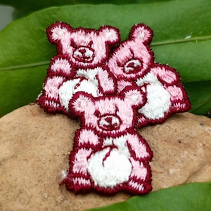May include: Three pink and white embroidered teddy bear patches with maroon outlines. The patches are stacked on top of each other.
