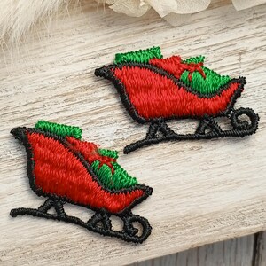 May include: Two embroidered Christmas sleigh patches. Each sleigh is red with green accents and a black outline. The sleighs are filled with gifts and have a black runner. The patches are on a light wood surface.