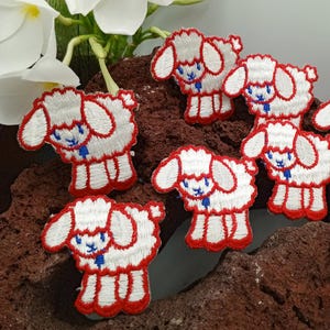 May include: Six white embroidered iron-on patches of lambs with red outlines. The lambs have blue eyes and a blue ribbon around their necks.