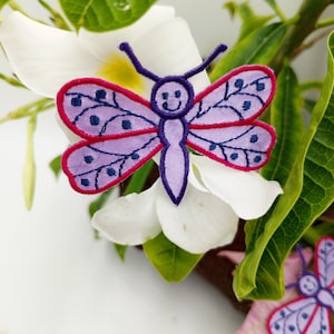 May include: Purple and red embroidered butterfly patch with a smiling face. The patch is on a white flower with green leaves.