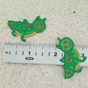 May include: Two green embroidered iron-on patches of smiling grasshoppers. The patches are about 1 inch in length.