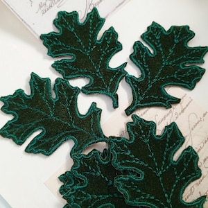 May include: Five dark green felt oak leaves with a light green embroidered outline. The leaves are arranged in a cluster on a white surface.