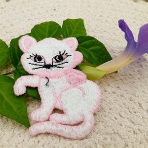 May include: White embroidered cat applique with pink accents. The cat has black eyes and a pink nose. The applique is perfect for adding a touch of whimsy to any project.