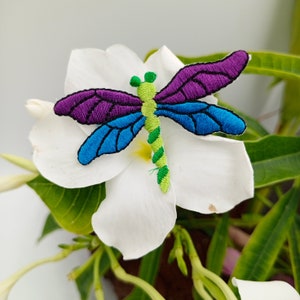 May include: A colorful embroidered dragonfly with purple, blue, and green accents is perched on a white flower.