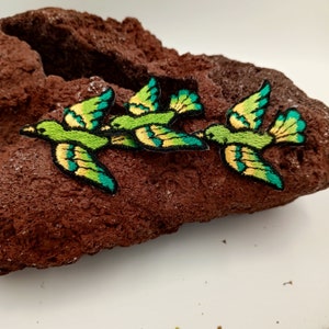 May include: Three green and yellow embroidered bird patches with teal accents. The patches are arranged in a row on a brown textured surface.
