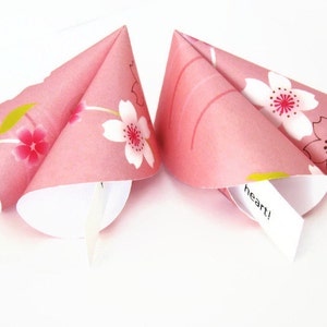 May include: Two pink fortune cookies with a floral cherry blossom pattern. The cookies are open, revealing white paper fortunes. One fortune reads "heart!"