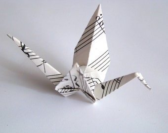 May include: A white origami crane, crafted from paper printed with musical notation. The crane is depicted with its wings spread, showcasing the intricate folds and the contrast of black notes against the white background. A symbol of peace and art.
