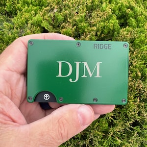 May include: A green metal wallet with the initials "DJM" engraved on the front. The word "RIDGE" is printed at the top. The wallet is held against a background of green moss.