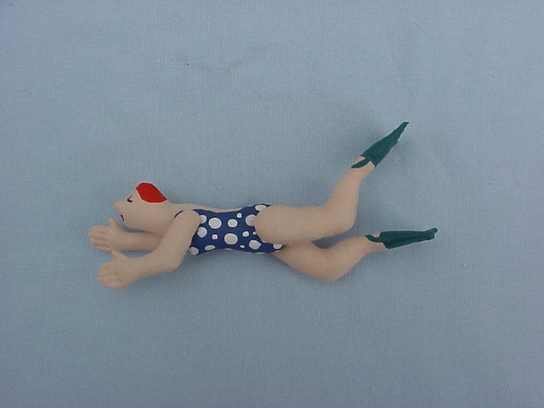 Op de afbeelding: Een handgemaakte pop van een zwemmer die een blauw-wit geblokt badpak en groene flippers draagt. De pop ligt op zijn zij met zijn armen uitgestrekt.