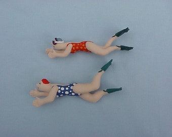 May include: Two handmade fabric dolls of swimmers wearing swimsuits and flippers. The doll on top is wearing a red and white swimsuit and the doll on the bottom is wearing a white and blue polka dot swimsuit.