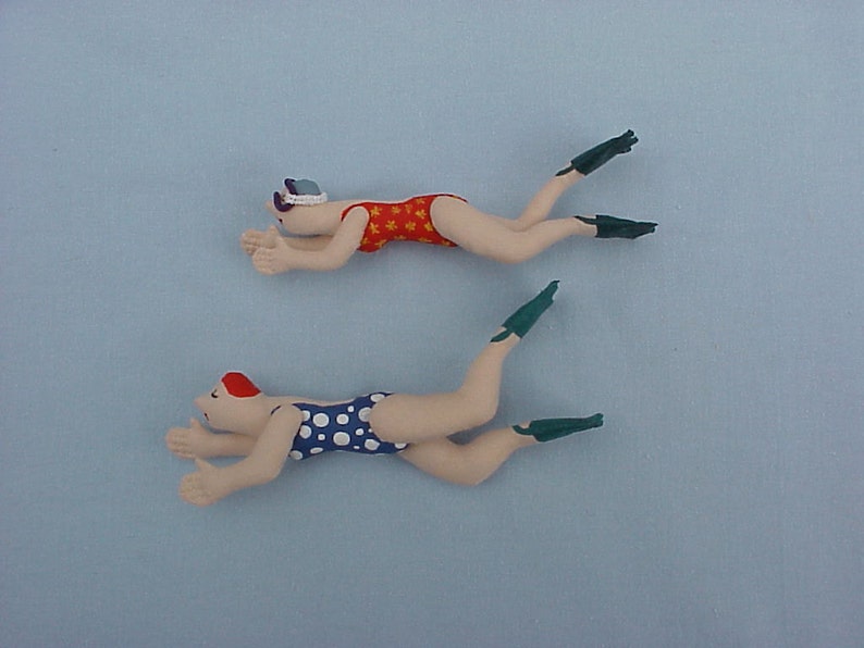 Op de afbeelding: Twee handgemaakte poppen van stof van zwemmers die badpakken en flippers dragen. De pop bovenaan draagt een rood-wit badpak en de pop onderaan draagt een wit-blauw gestippeld badpak.